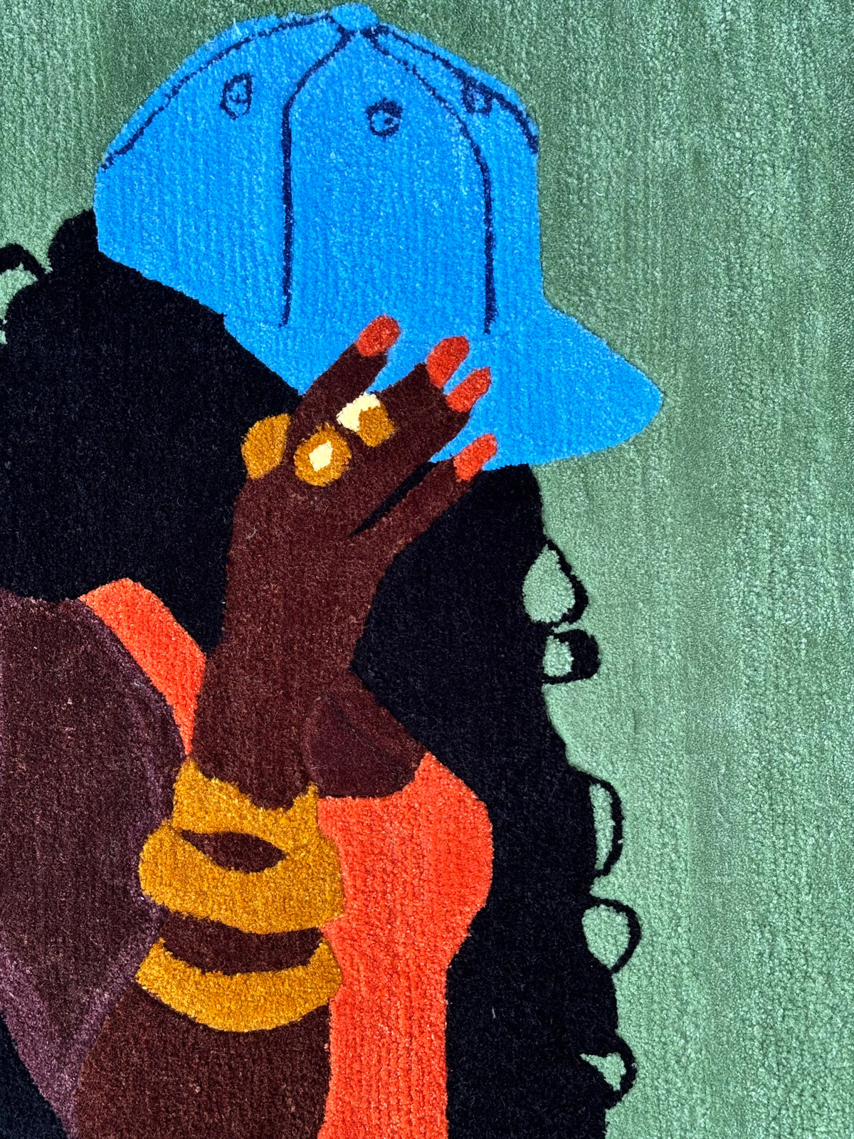 A rug showing a woman in profile wearing a bright blue cap, with dark curly hair, a coral-orange top, gold rings, and a green textured background.