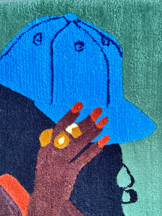 A close-up of a colorful textured rug featuring an illustration of a woman wearing a bright blue cap, with a dark hand in the foreground and a green background.